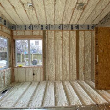 Interior of a spray-foam insulated room with exposed studs, window, and unfinished floor and ceiling