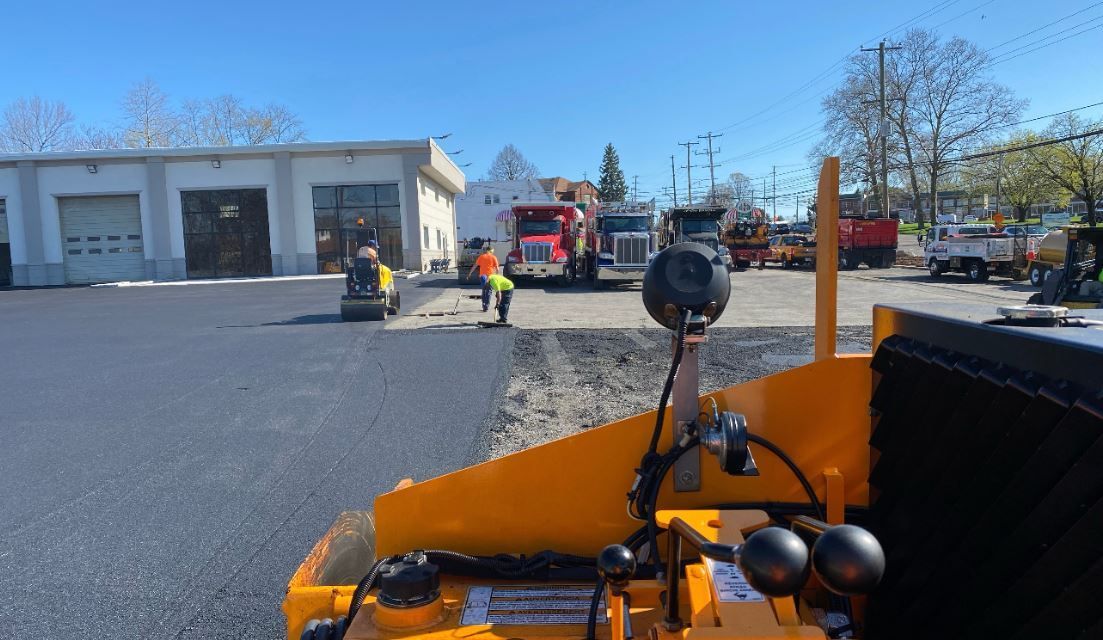 A yellow asphalt roller is being used to spread asphalt in a parking lot.