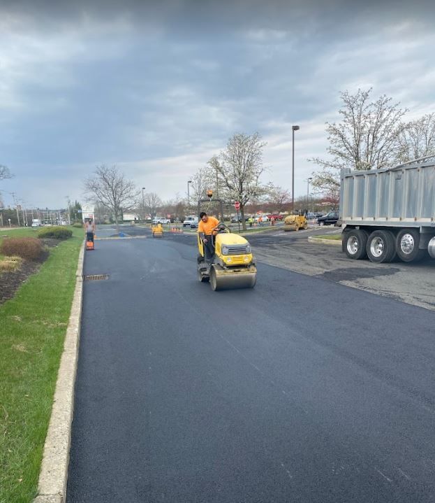 A man is driving a roller on a road next to a dump truck.