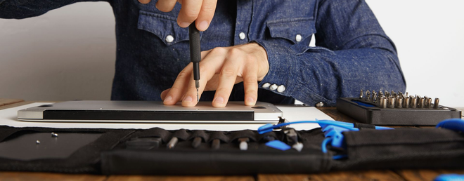 A person wearing a denim shirt uses a precision screwdriver to work on the bottom panel of a silver laptop.