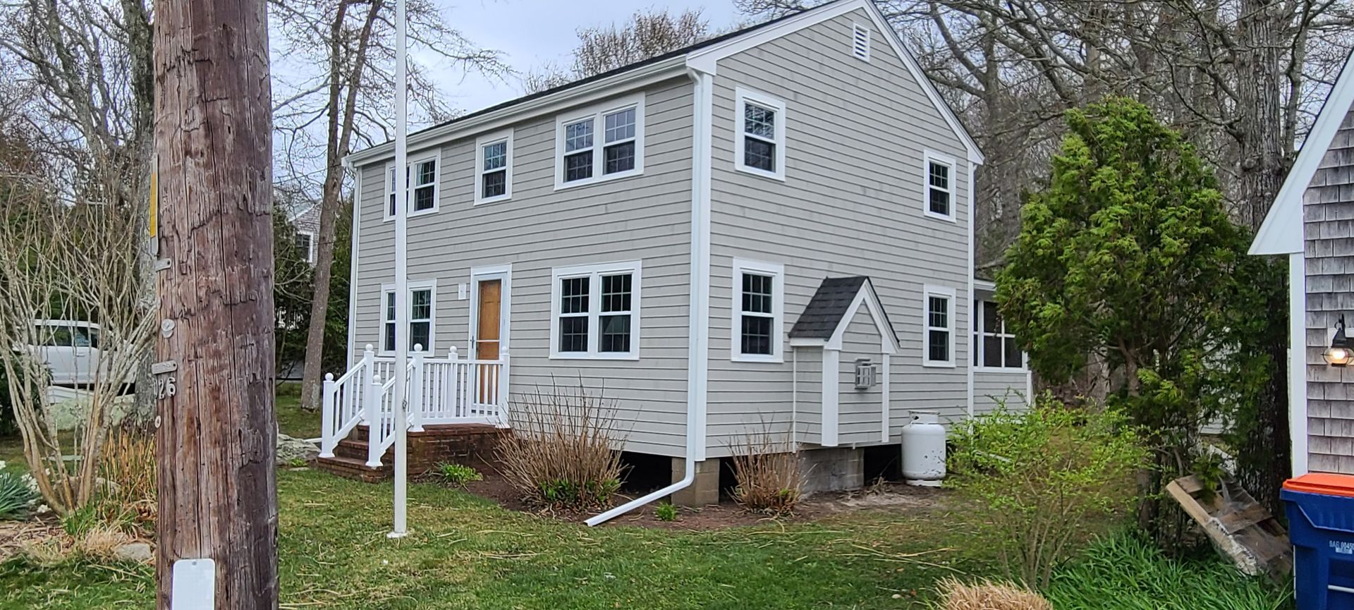 Two-story light gray house with dark roof and white trim; door with steps; surrounded by trees and grass.
