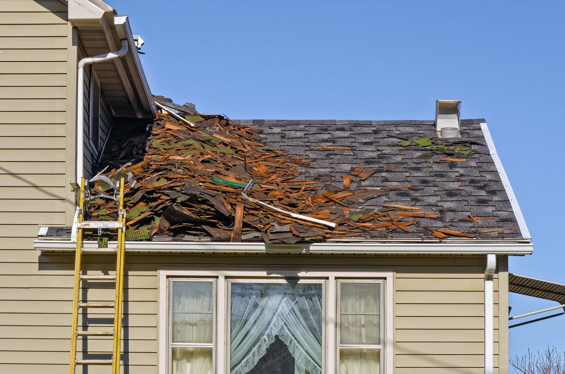 House with damaged roof and debris, sunlight, and a ladder against the side.