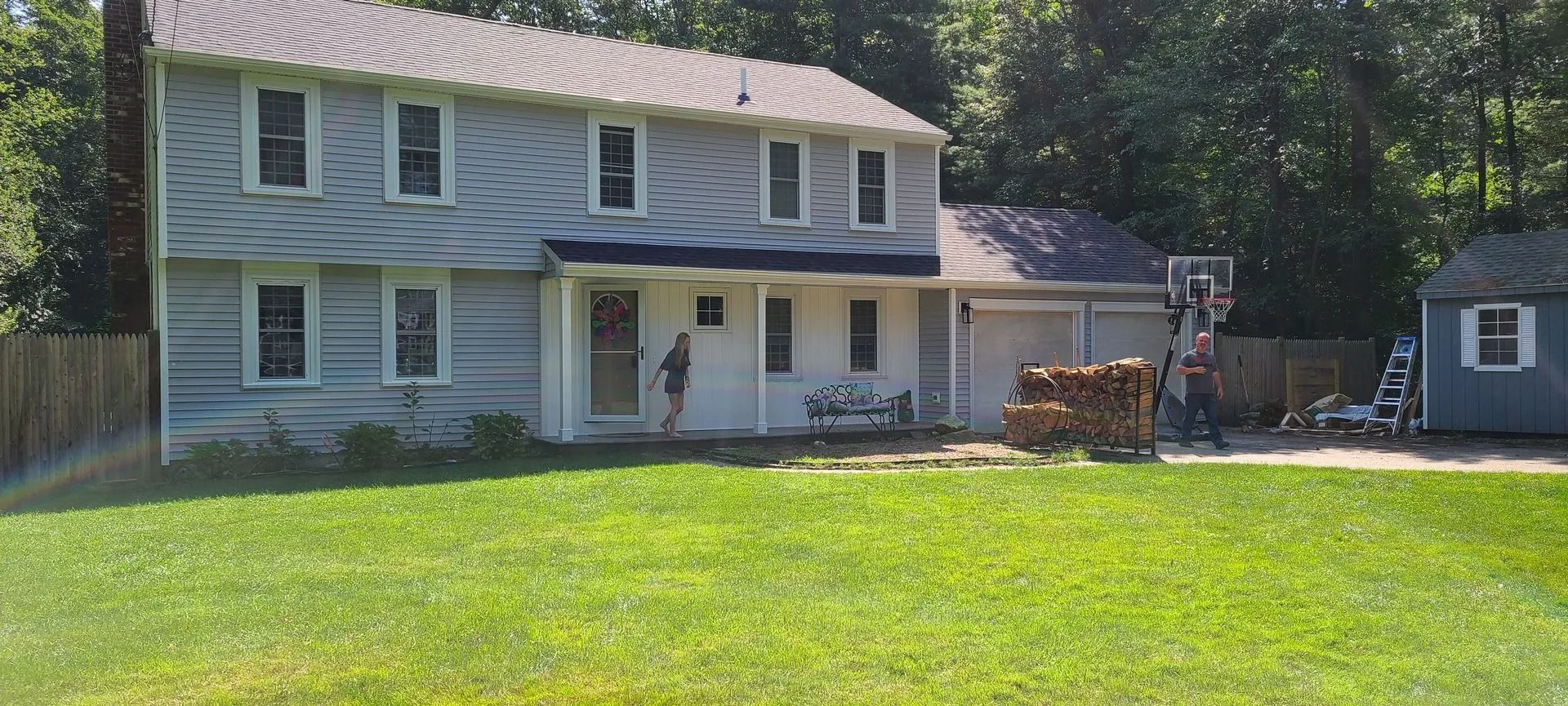 Two-story house with green lawn, trees, and a small outbuilding. A person stands near the front door.