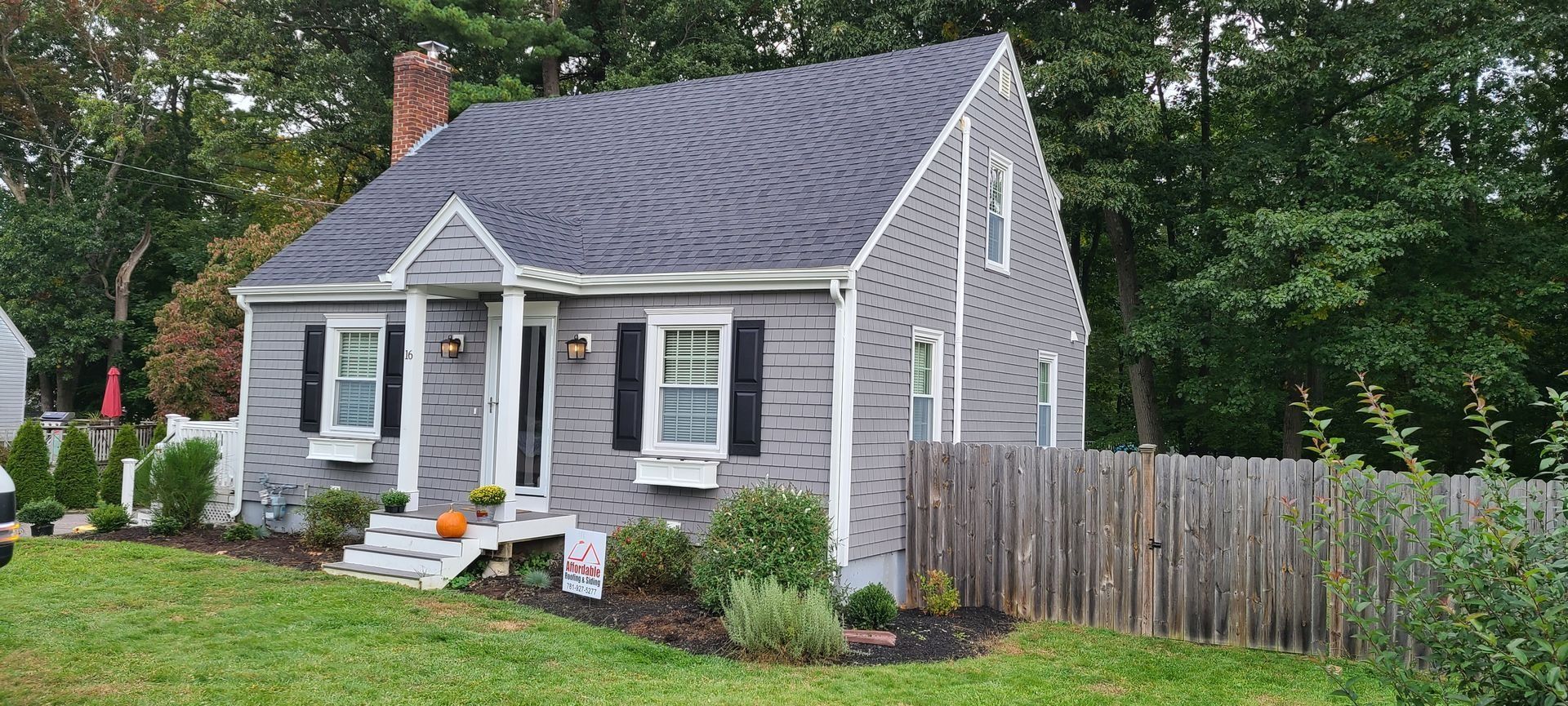 Gray house with black shutters, a wooden fence, and a brick chimney. A pumpkin sits on the porch.