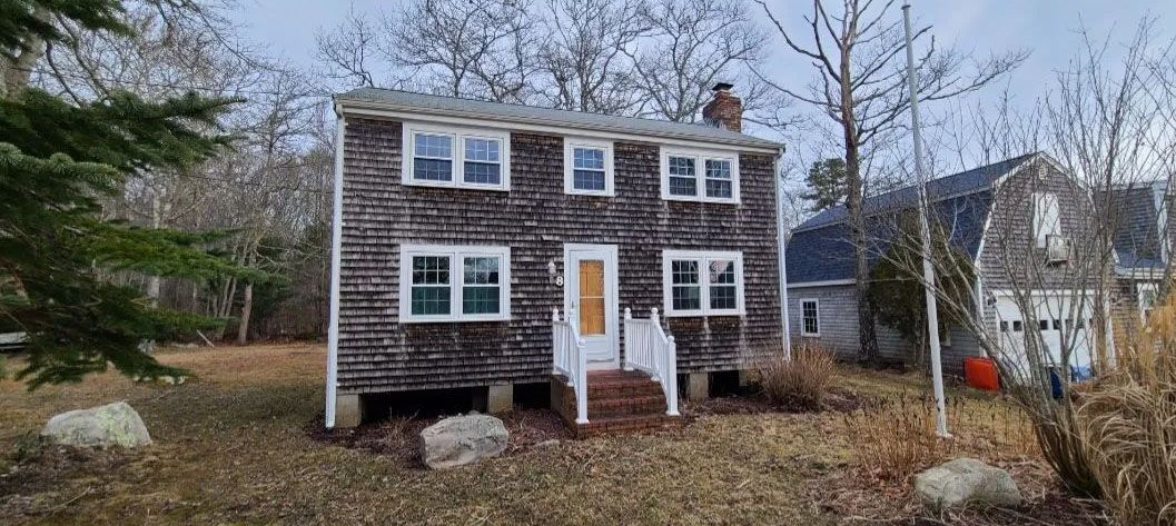 Two-story house with workers on roof. Blue sky with fluffy clouds. Green grass and trees surround the house.