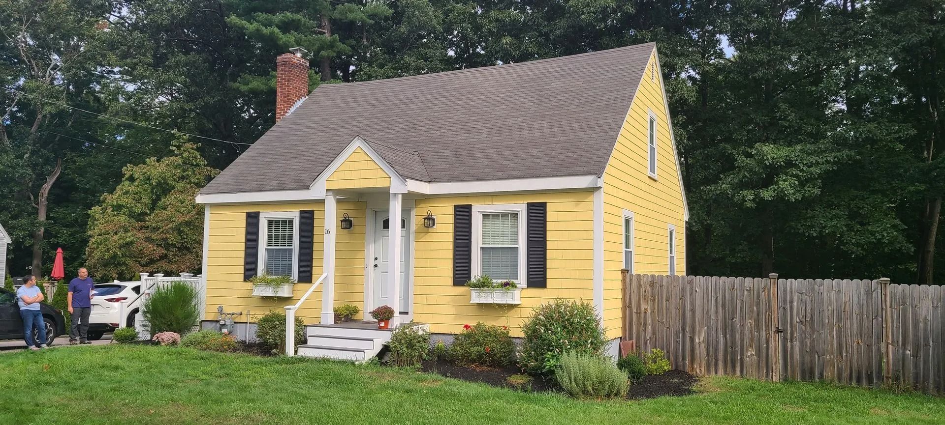 Yellow house with black shutters, brown roof, and wooden fence. Green lawn and trees.