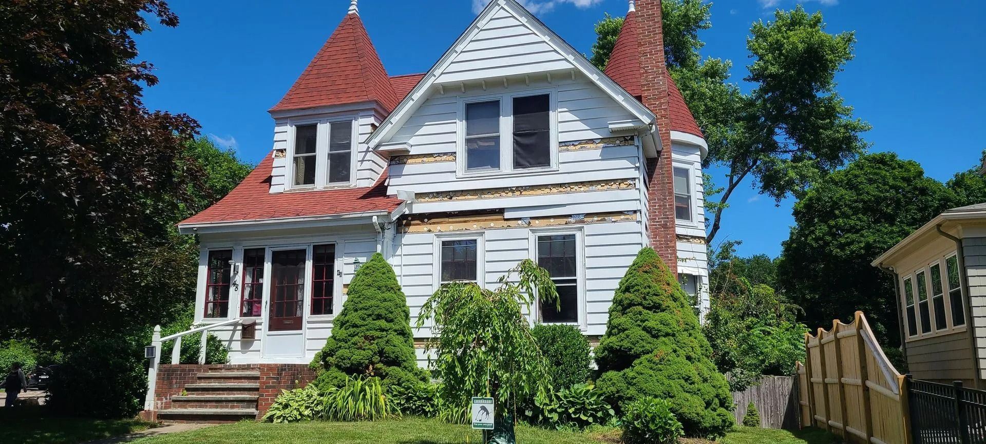 White Victorian house with red roof and turrets, surrounded by trees and green grass on a sunny day.