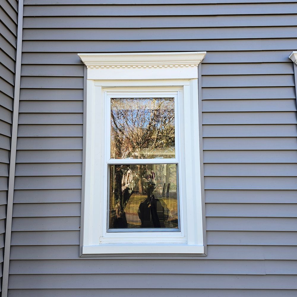 White-framed window with decorative molding on a gray siding wall reflecting trees and a person.