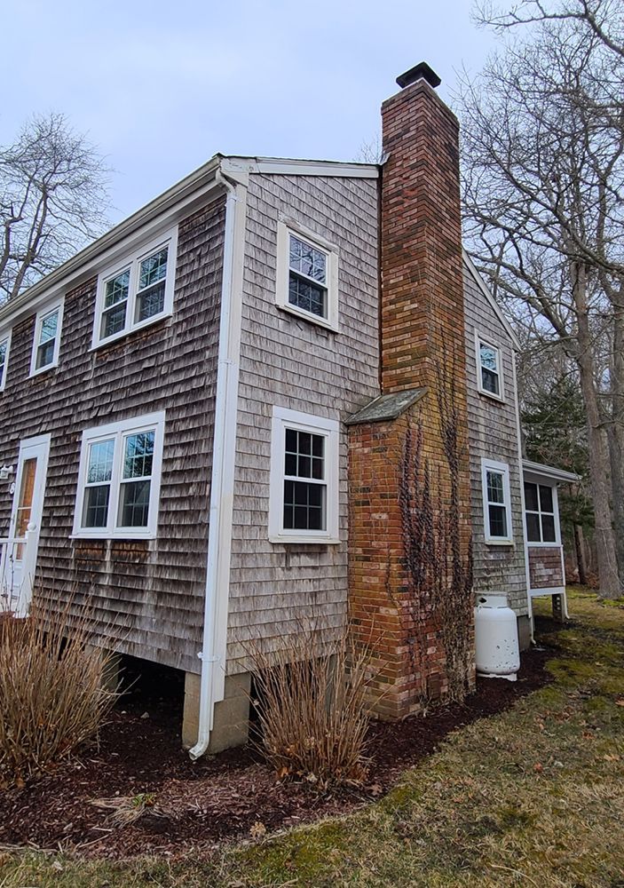 Two-story weathered shingle house with a brick chimney. White windows, brown mulch, and overcast sky.
