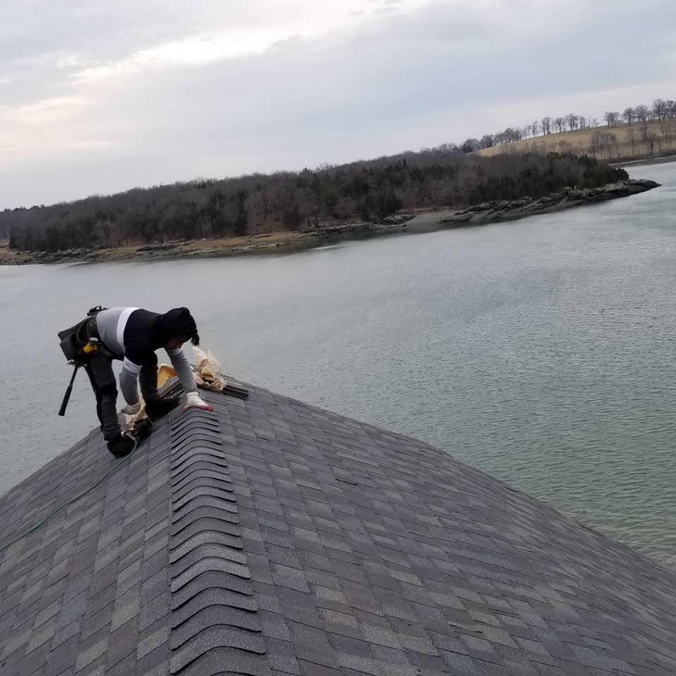 Two people work on a roof near water. One kneels with tools; the other handles roofing material. Overcast day.