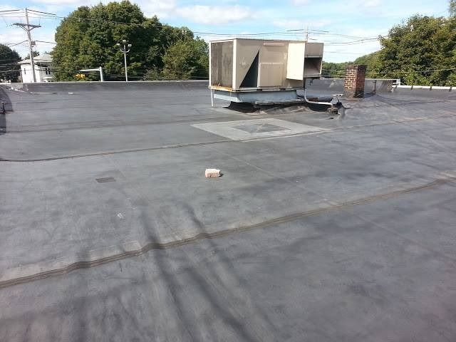 Flat, dark roof with HVAC unit and brick chimney against a treeline under a bright sky.
