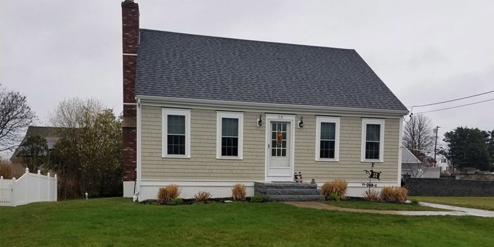 A one-story beige house with a brick chimney and a gray roof on a cloudy day.