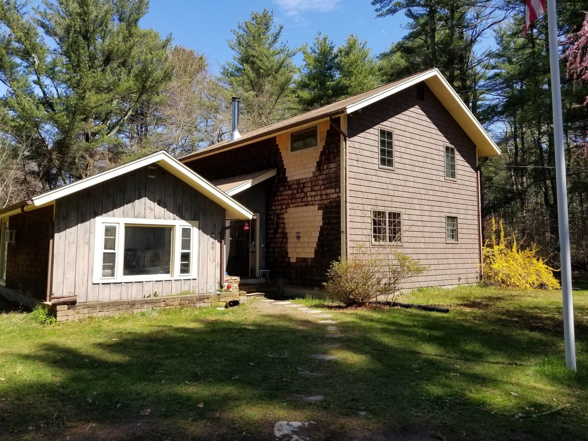 Brown house with an attached shed, surrounded by trees and greenery under a blue sky.