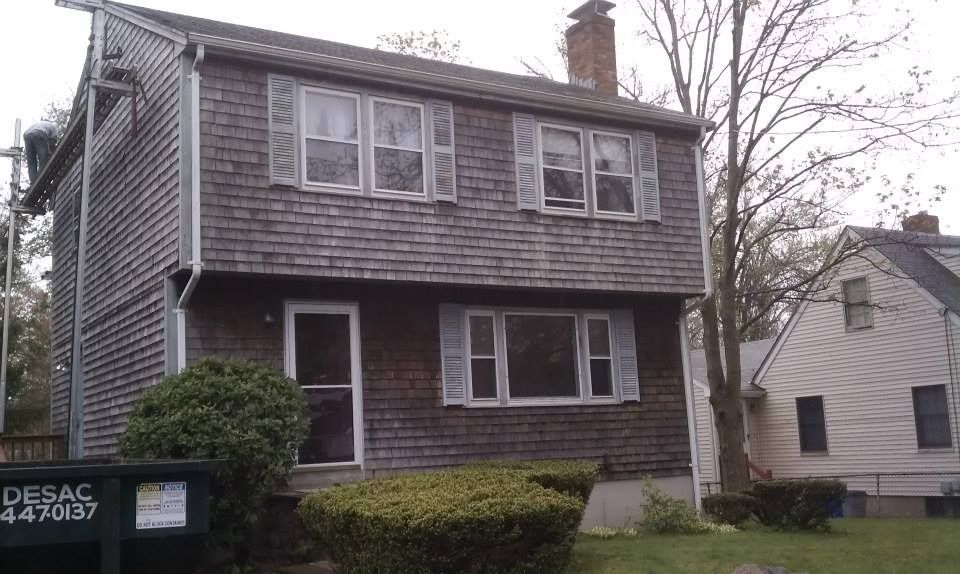 Two-story house with weathered cedar siding, white trim, and a small front yard with shrubs and a dumpster.
