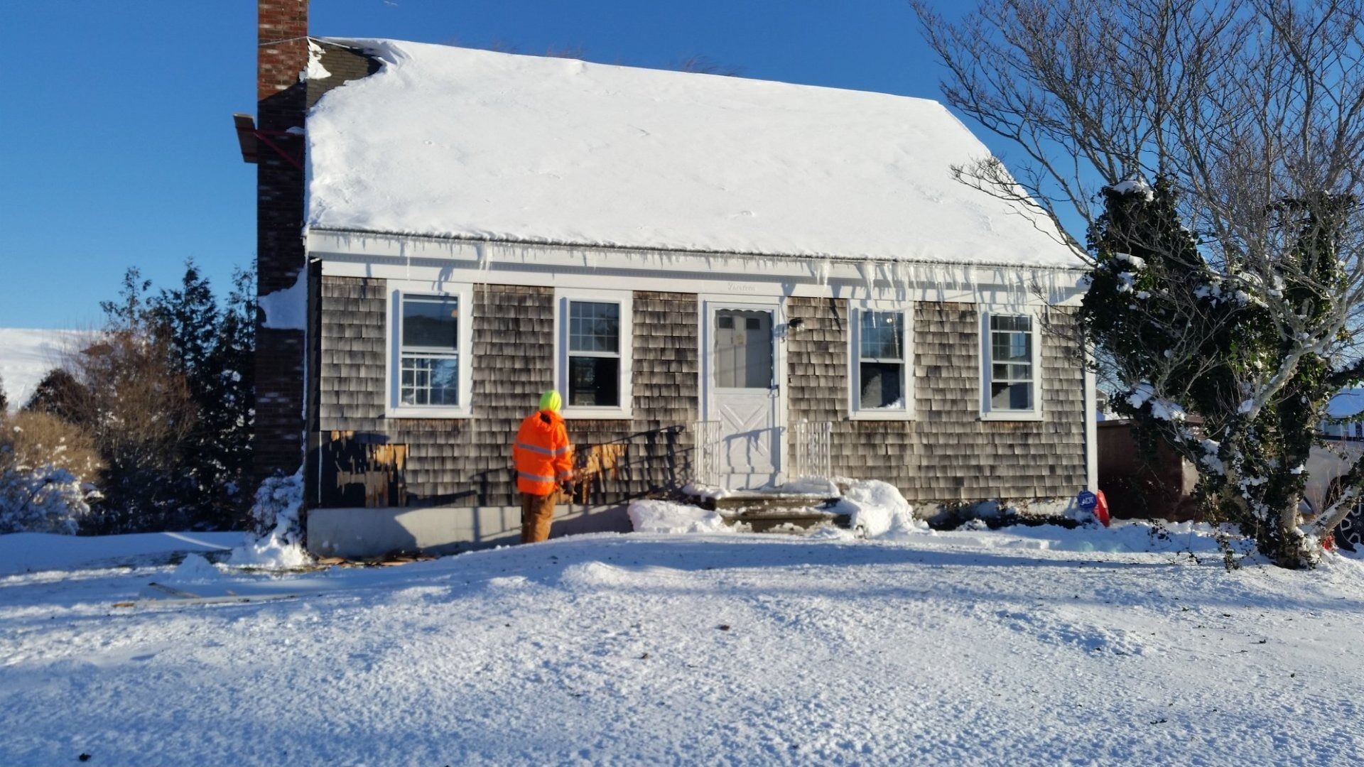 Snow-covered cottage with icicles, person in orange jacket, sunny winter day.