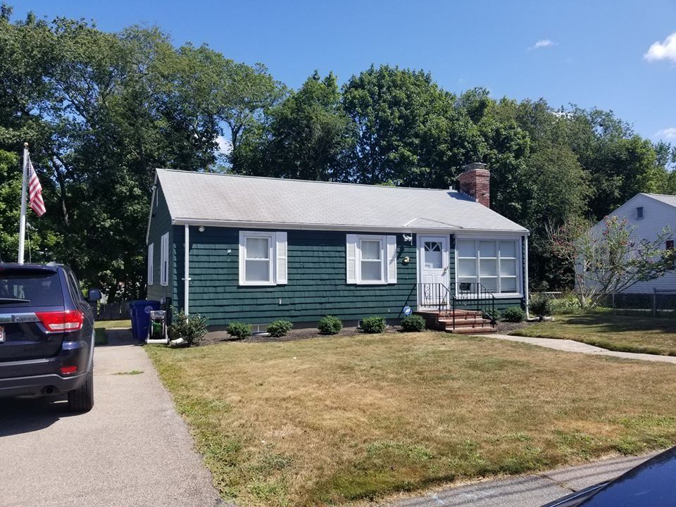 Green house with white trim, American flag, driveway, brown lawn, blue sky.