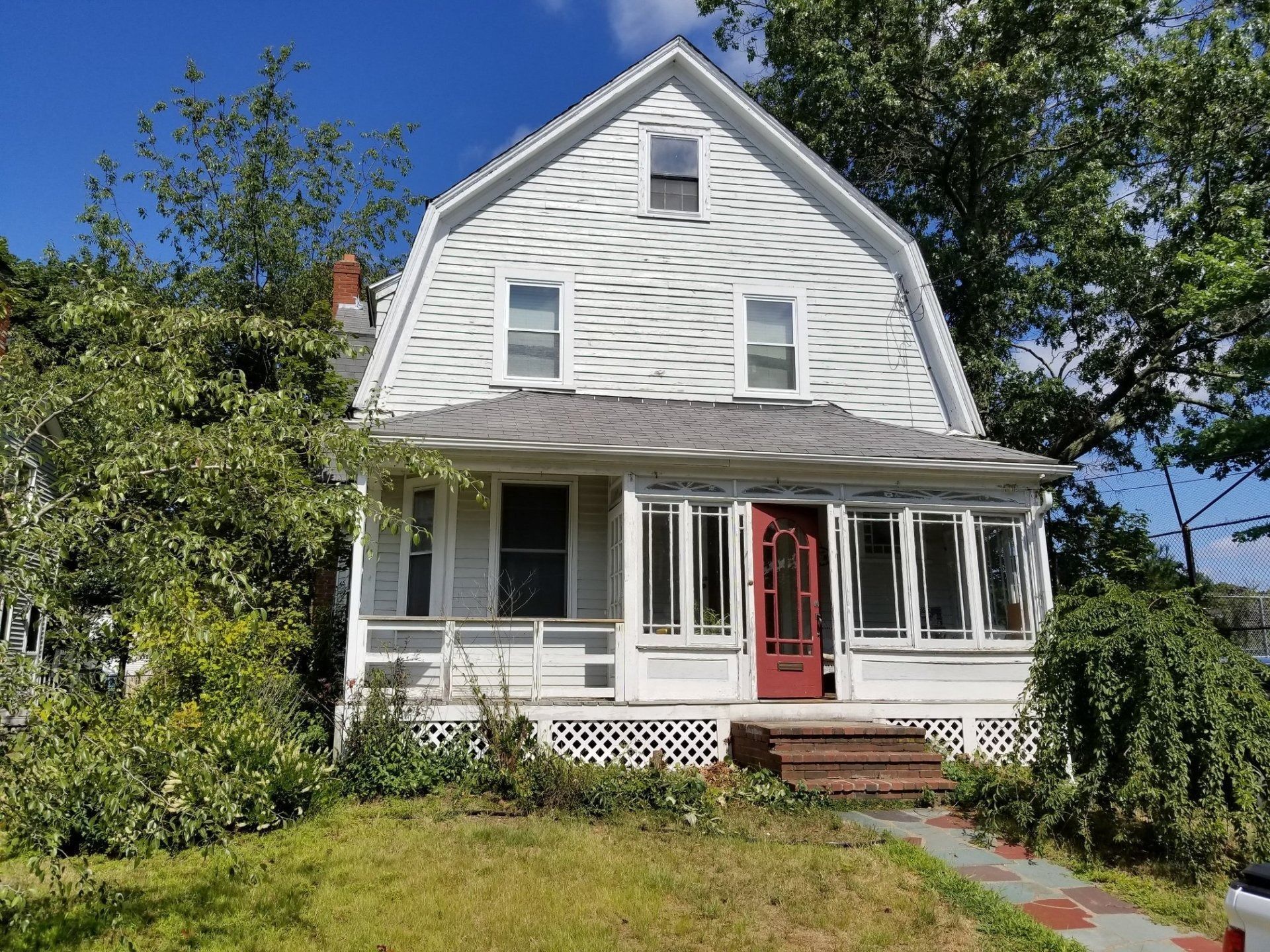 White house with a Dutch gambrel roof, porch, red door. Overgrown yard.