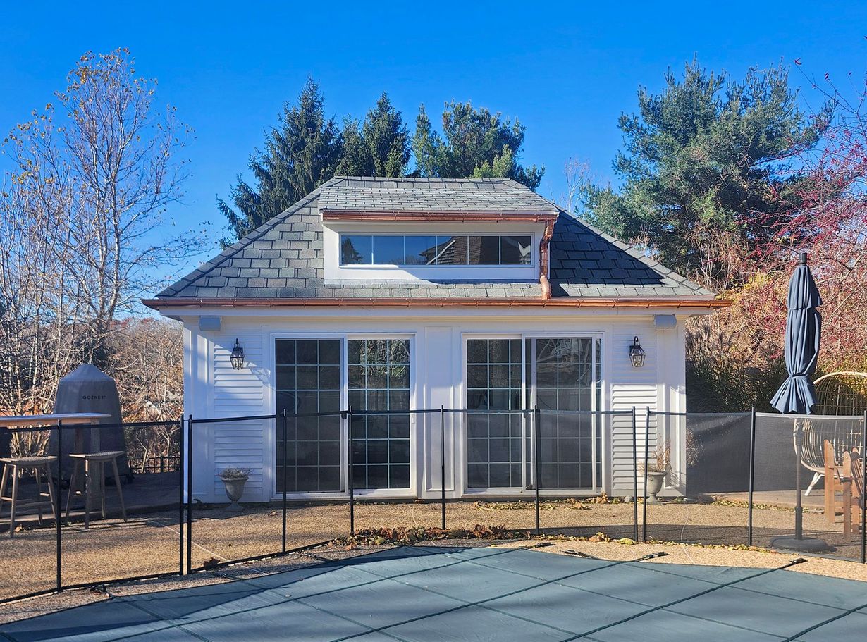 Triangular roof with grey shingles, small windows, and large wooden-framed windows on a white building.