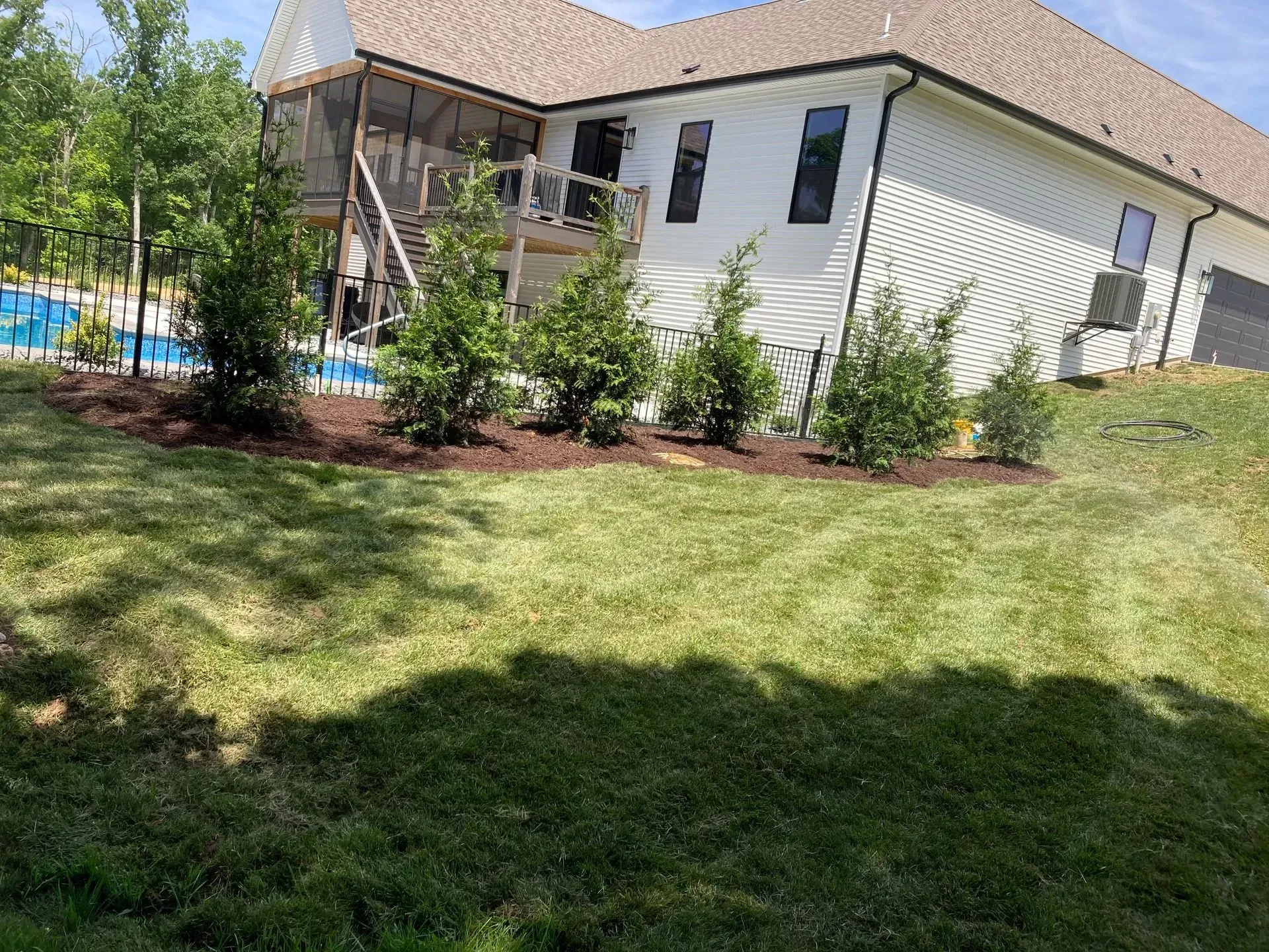 A white house with a screened porch and a row of shrubs along the side. Green lawn slopes downward.