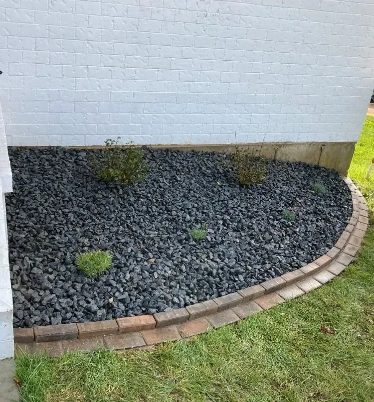 A landscaped garden bed with dark grey stone and small plants, bordered by a brick edge next to a white brick wall.