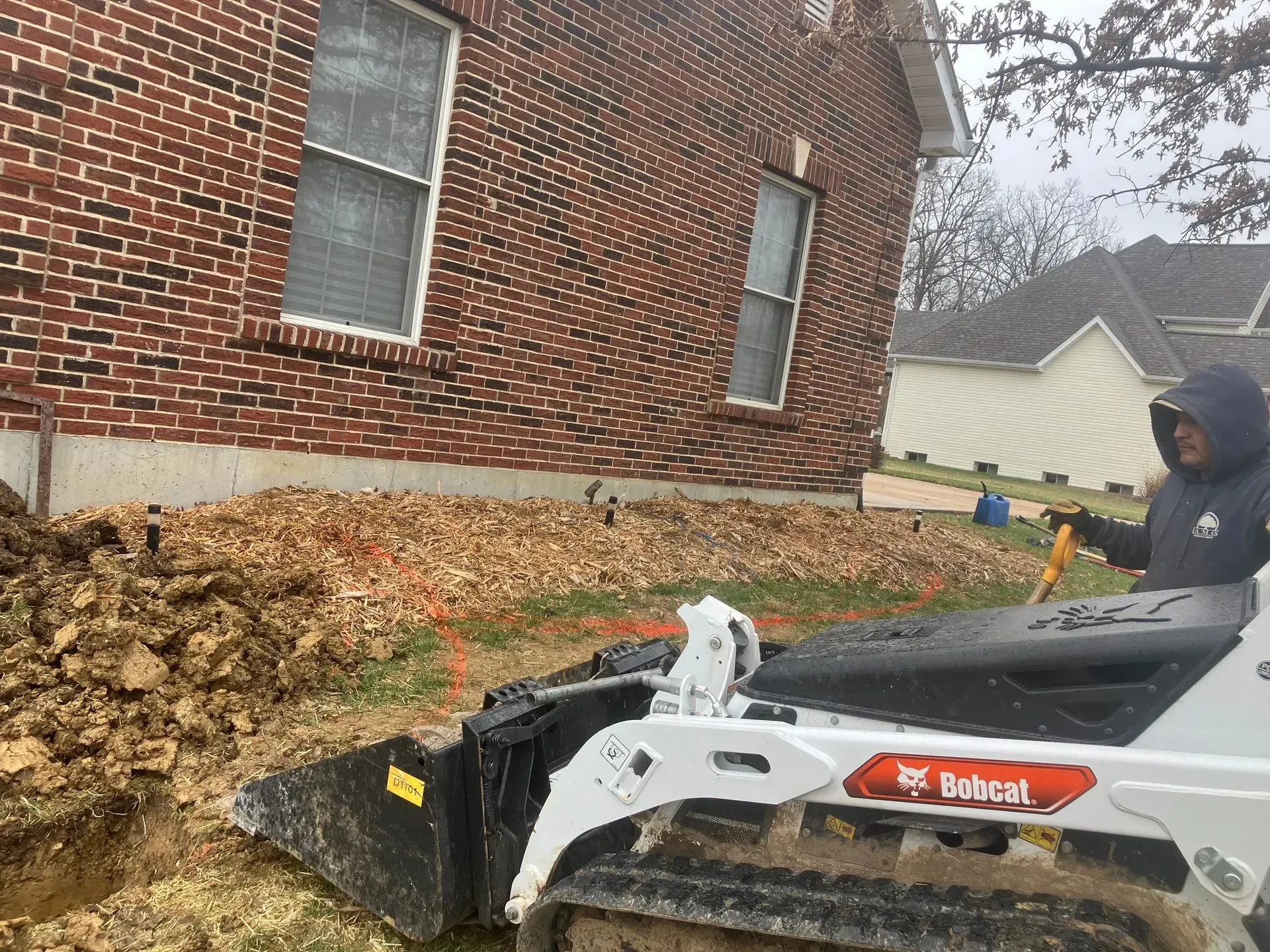 A Bobcat skid-steer loader excavates soil near a brick building. A person operates the machine on a cloudy day.