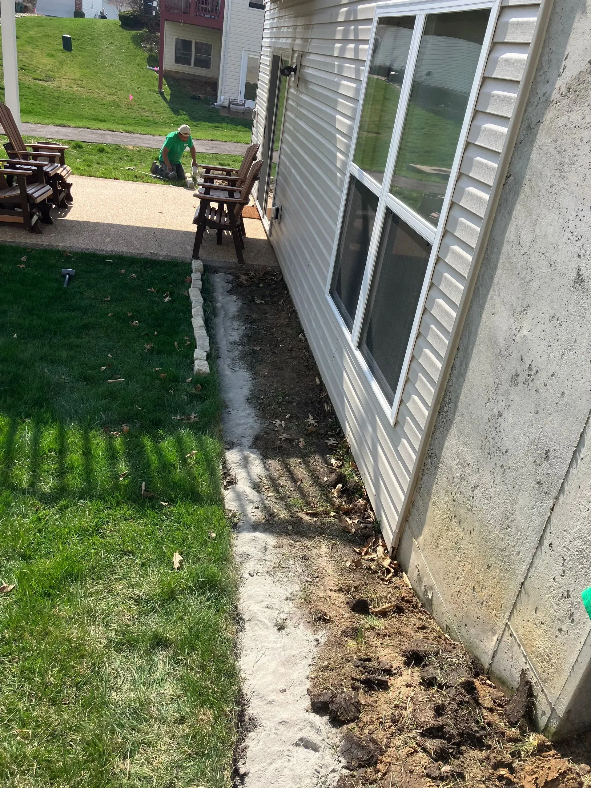 Side of a house with windows, next to a narrow patch of dirt and grass. Someone works in the background.