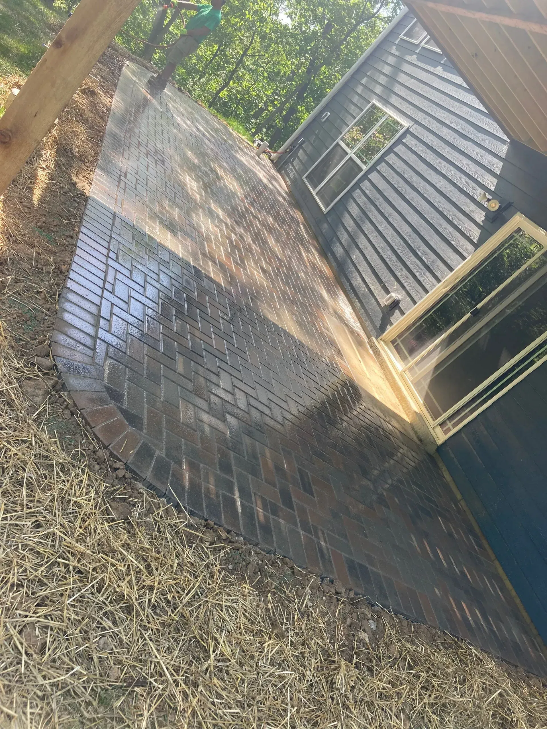 A dark brick patio curves alongside a blue-gray shed with two windows, surrounded by gravel and trees.