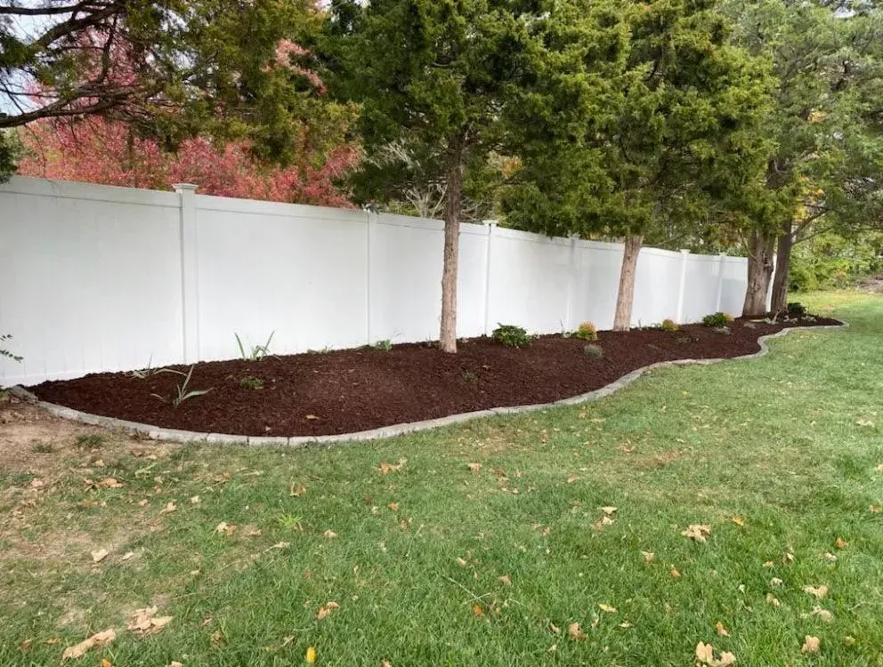 White fence borders a mulched garden bed with trees; green lawn in the foreground.