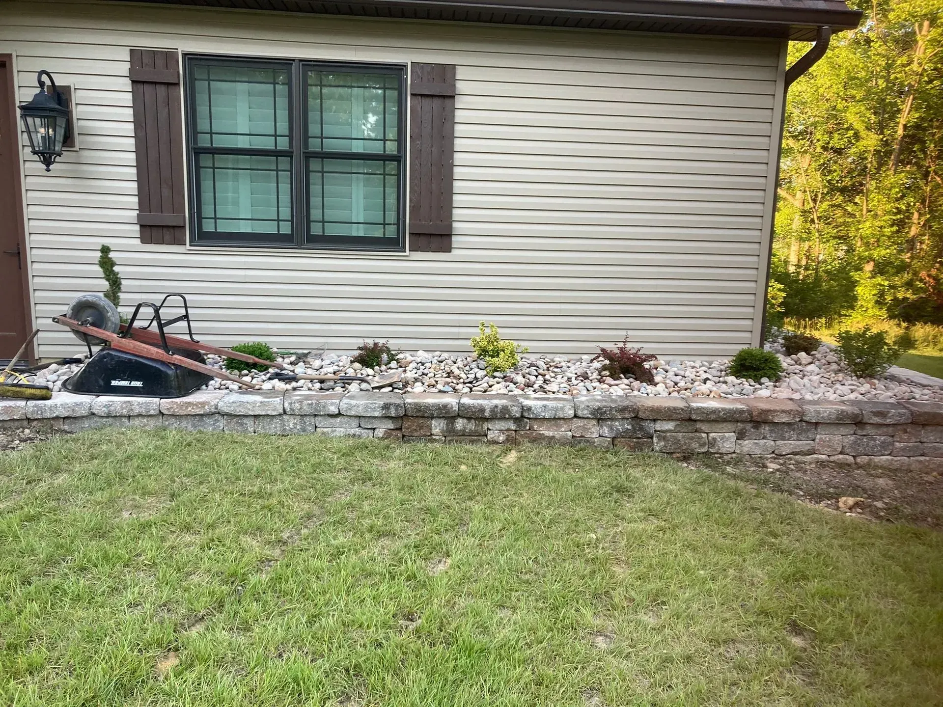 A house exterior with a small rock garden border. Beige siding, brown shutters, and green lawn.