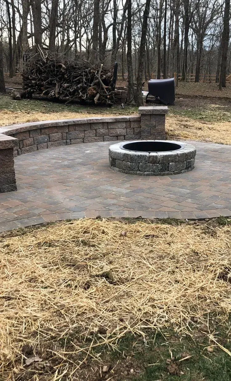 Stone fire pit and patio area with retaining wall, surrounded by dry grass and trees.