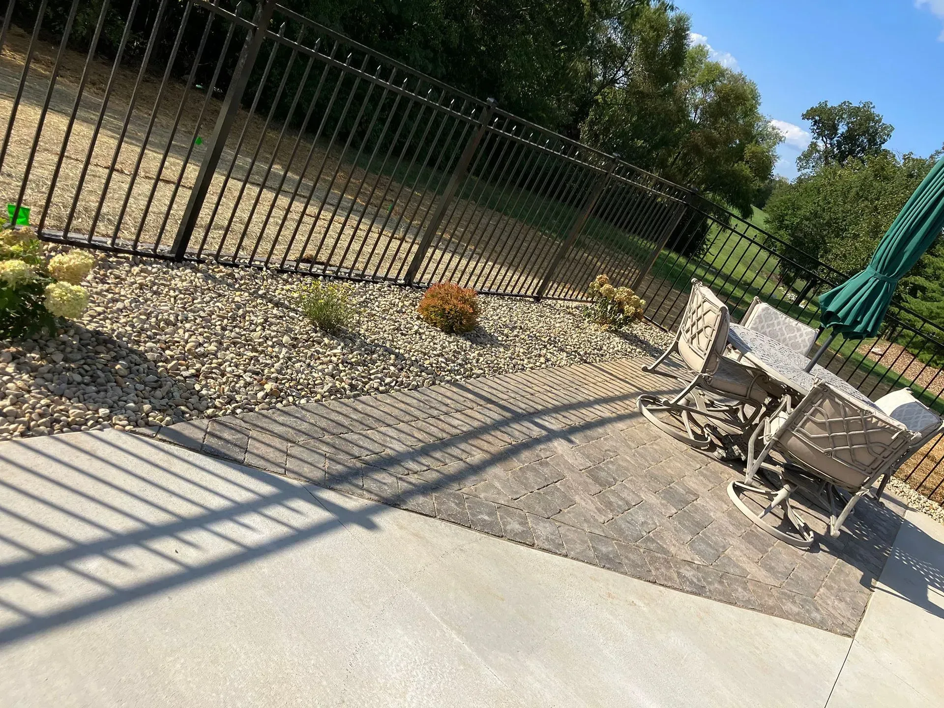 Patio with seating, gravel bed, black fence, and trees under a blue sky.