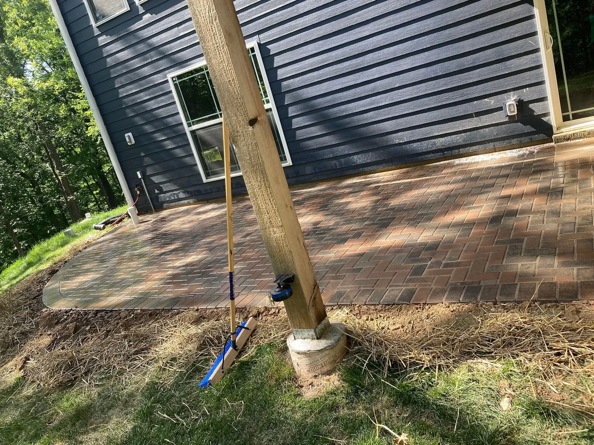 Wooden support post on brick patio next to a dark blue house. A broom leans against the post.