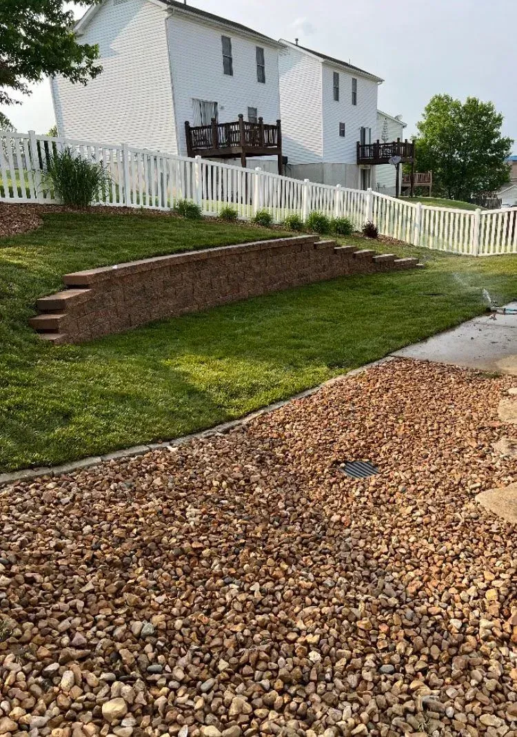A backyard with a tiered lawn, retaining wall, and white fence. Sprinkler in operation.
