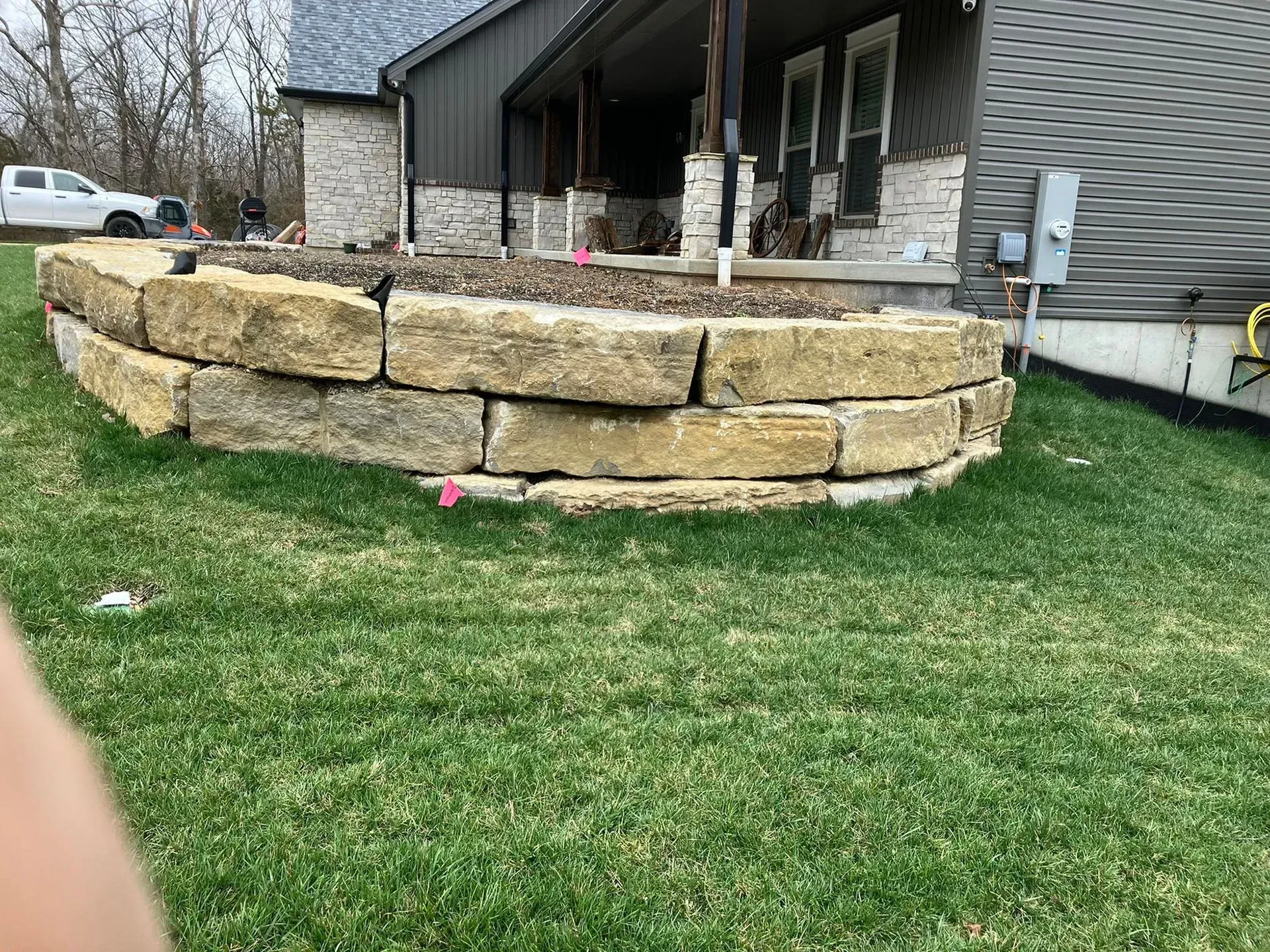 Stone retaining wall in front of a house, with green grass in the foreground and a porch in the background.