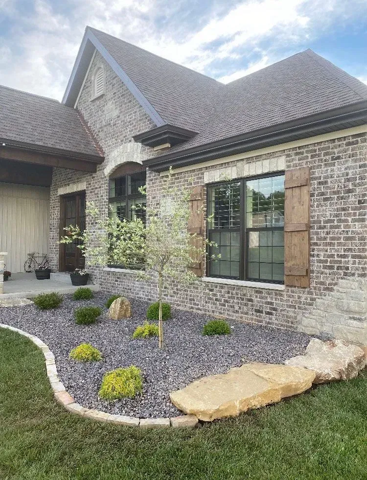 Exterior of a brick house with landscaping. Features dark shutters, windows, stone border, and rock accents.