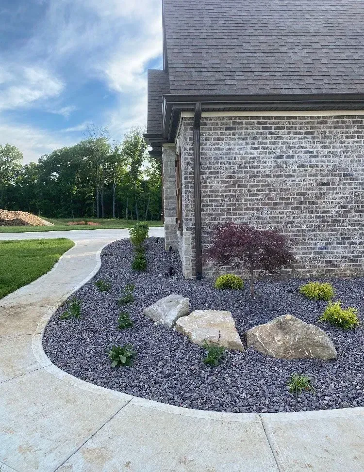 Landscaped front yard with dark stone, boulders, and a curved sidewalk next to a brick house.