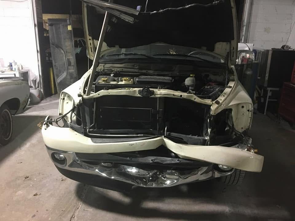 Severely damaged white car front with hood open in a garage, showing exposed engine and crumpled bumper.