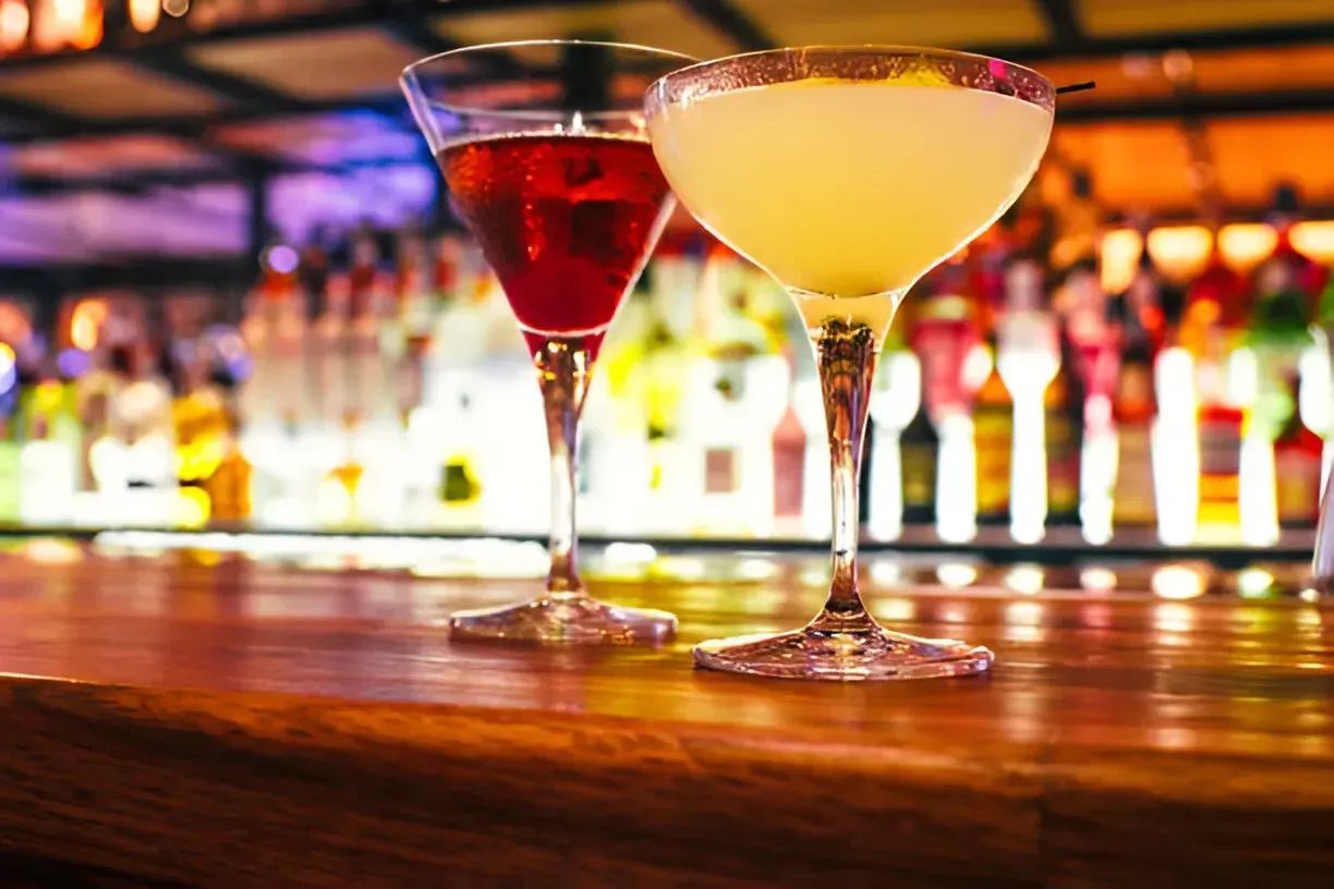 Two colorful cocktails in martini glasses on a bar counter with blurred bottles in the background