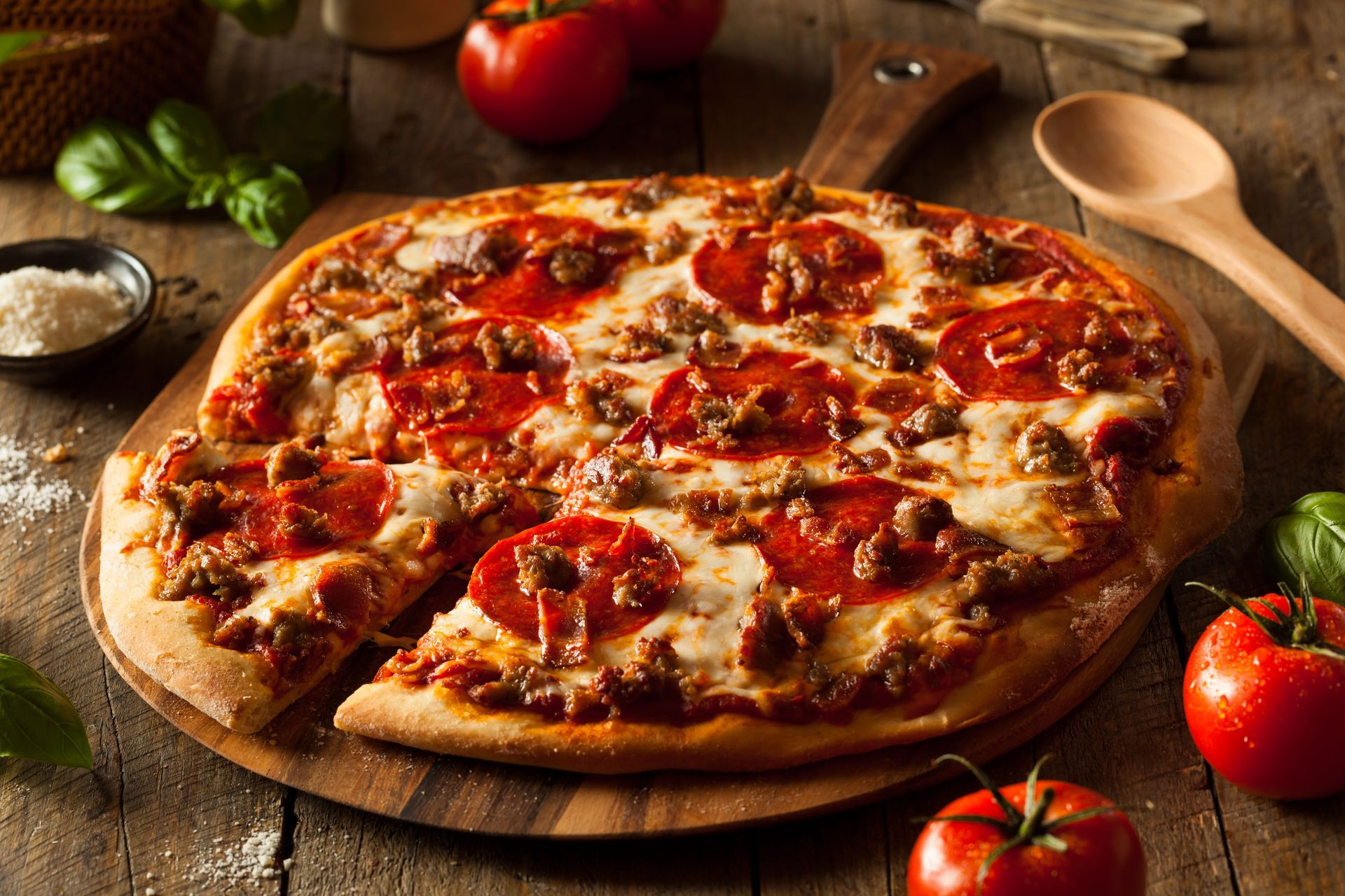 Fresh pepperoni pizza on a wooden board with basil, tomatoes, and a spoon on a rustic table