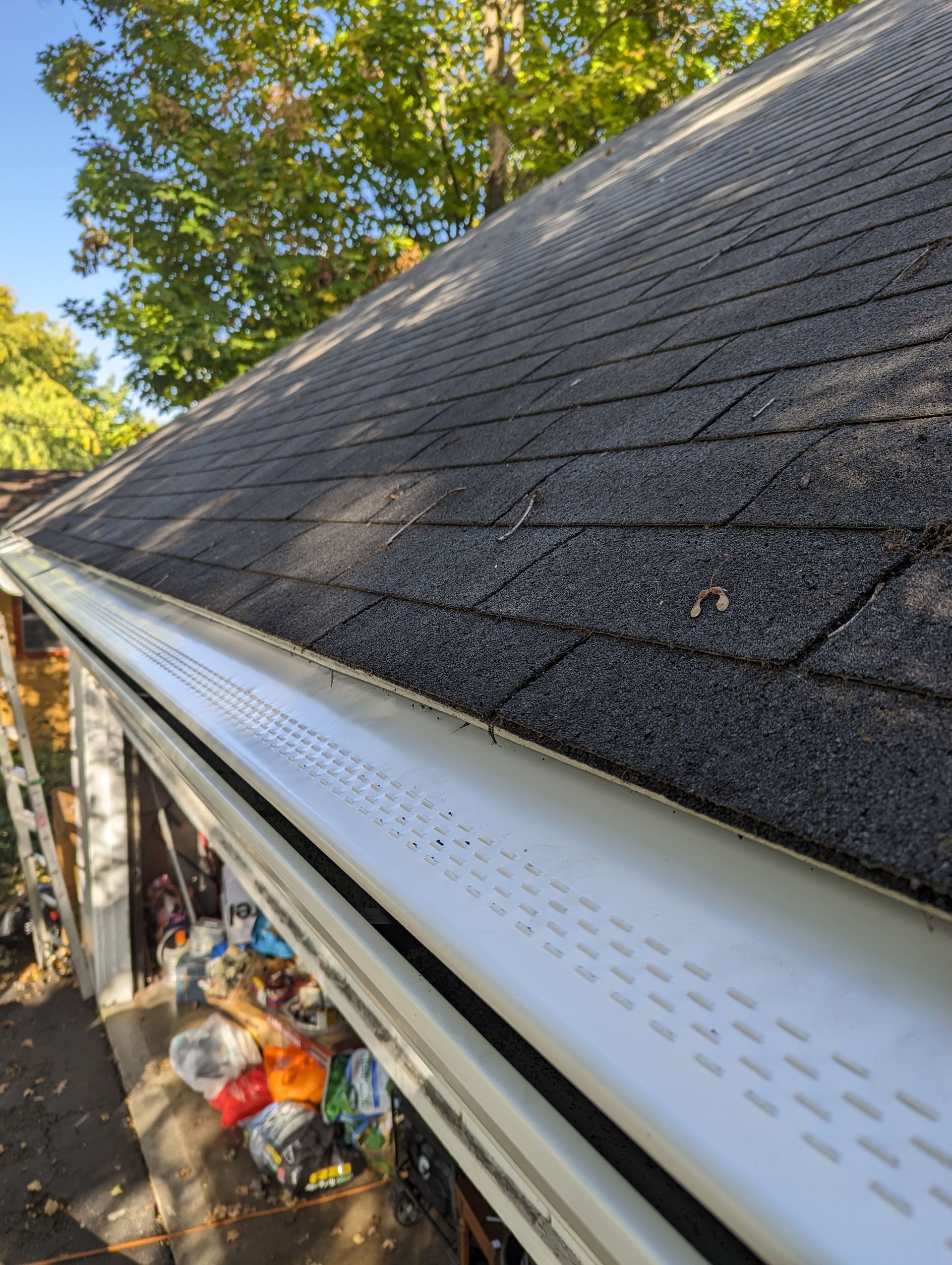 Close-up of a roof with black shingles and a white gutter with a leaf guard. Outdoors with trees in the background.
