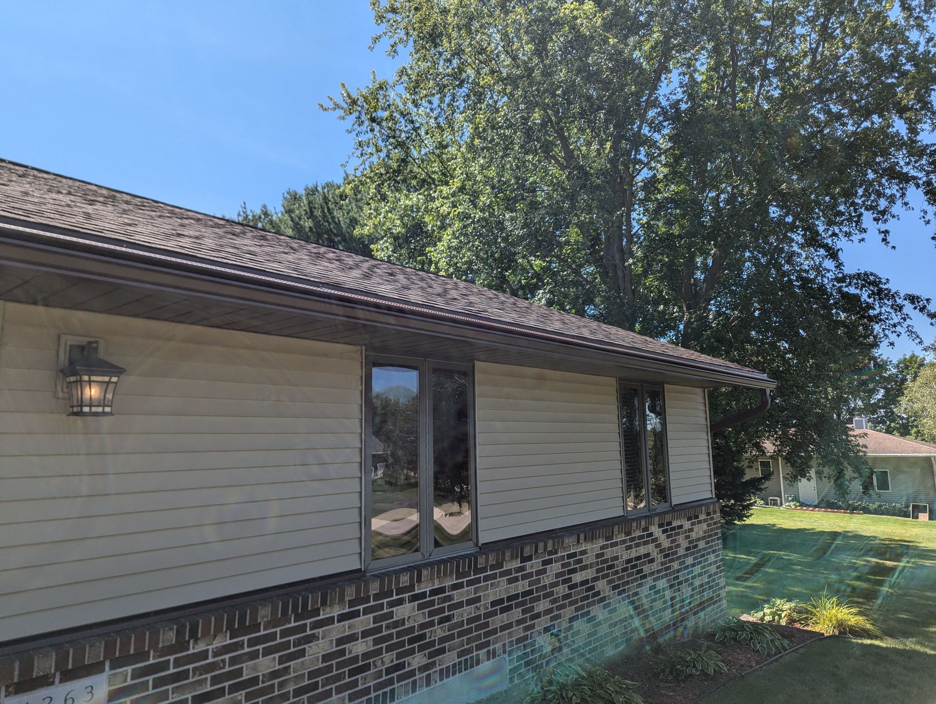 Tan house with brown trim and roof, brick base, windows, and a tree, outdoors on a sunny day.