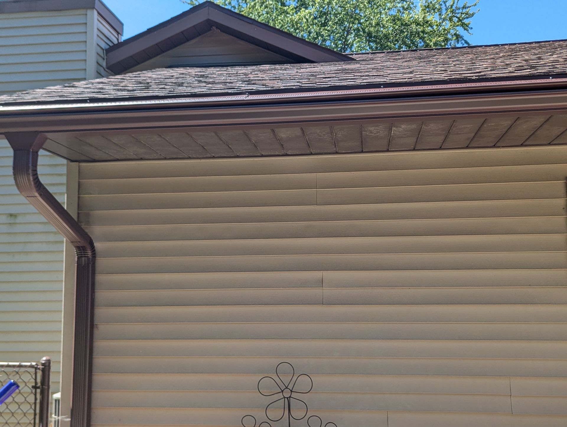 Tan vinyl siding on a house with brown gutters and roof. Sunny day.