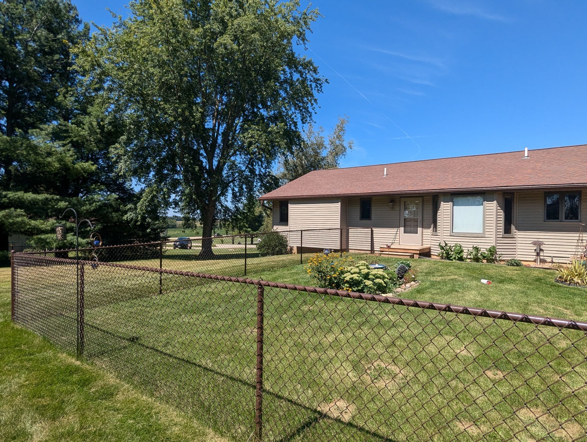 Brown chain-link fence surrounds a house on a sunny day; trees in the background.
