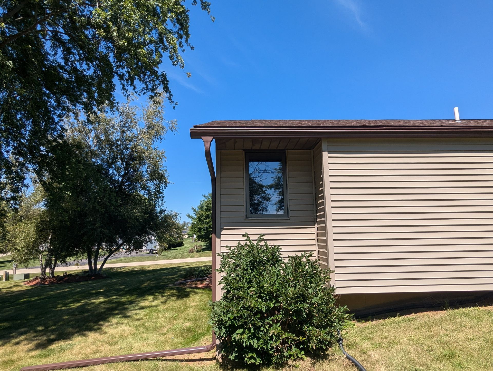 Side of a beige building with a small window, brown trim, and a bush in front, under a clear blue sky.