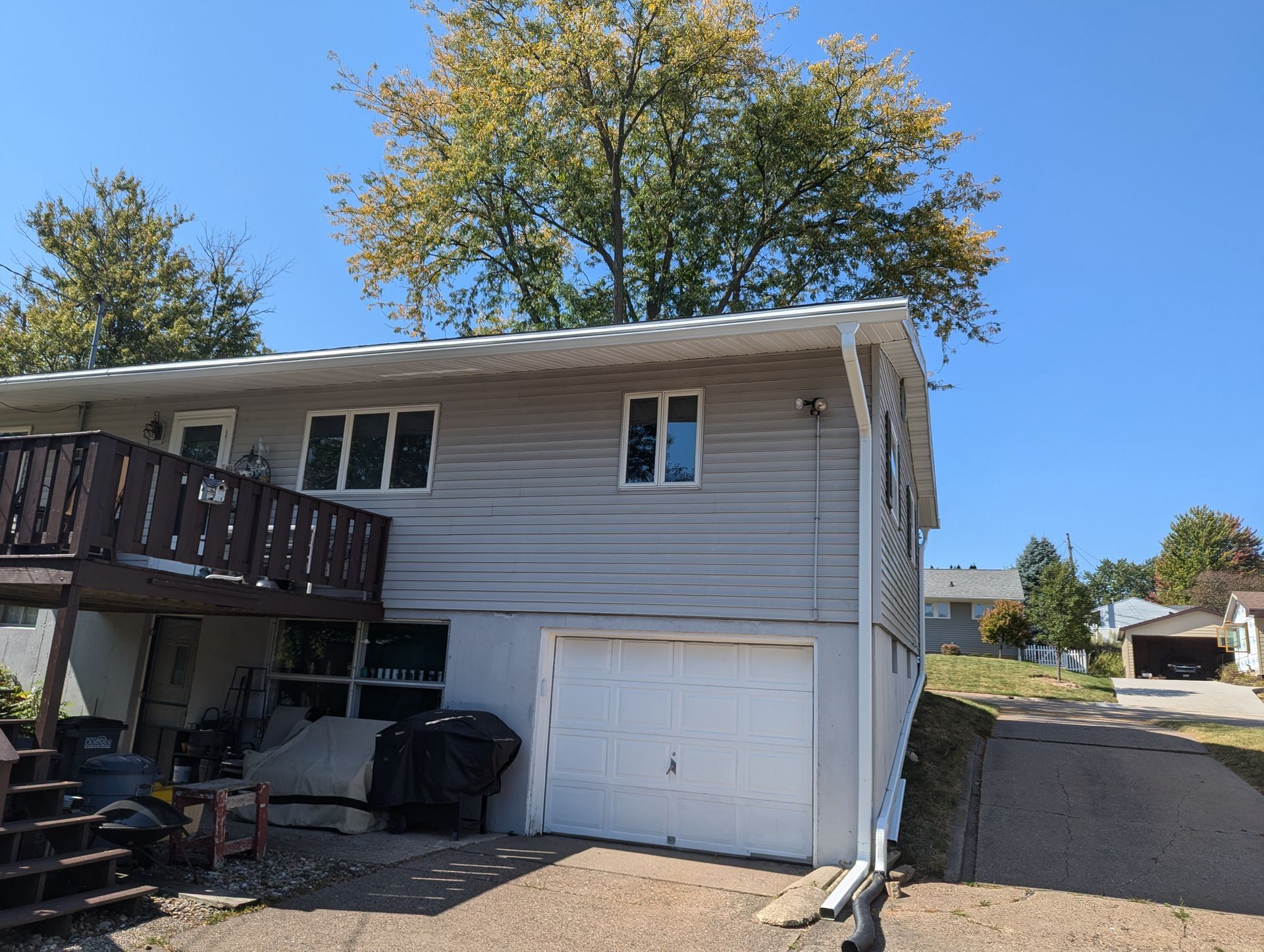 Two-story house with gray siding, white garage door, and wooden balcony. A tree shades the roof.