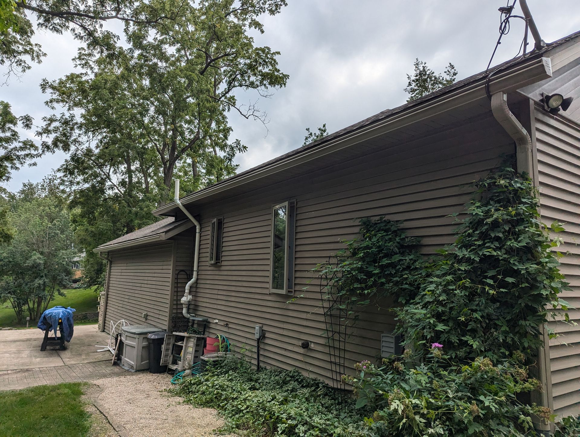 Brown house with siding, gutters, and a climbing vine. A man works near the driveway under an overcast sky.