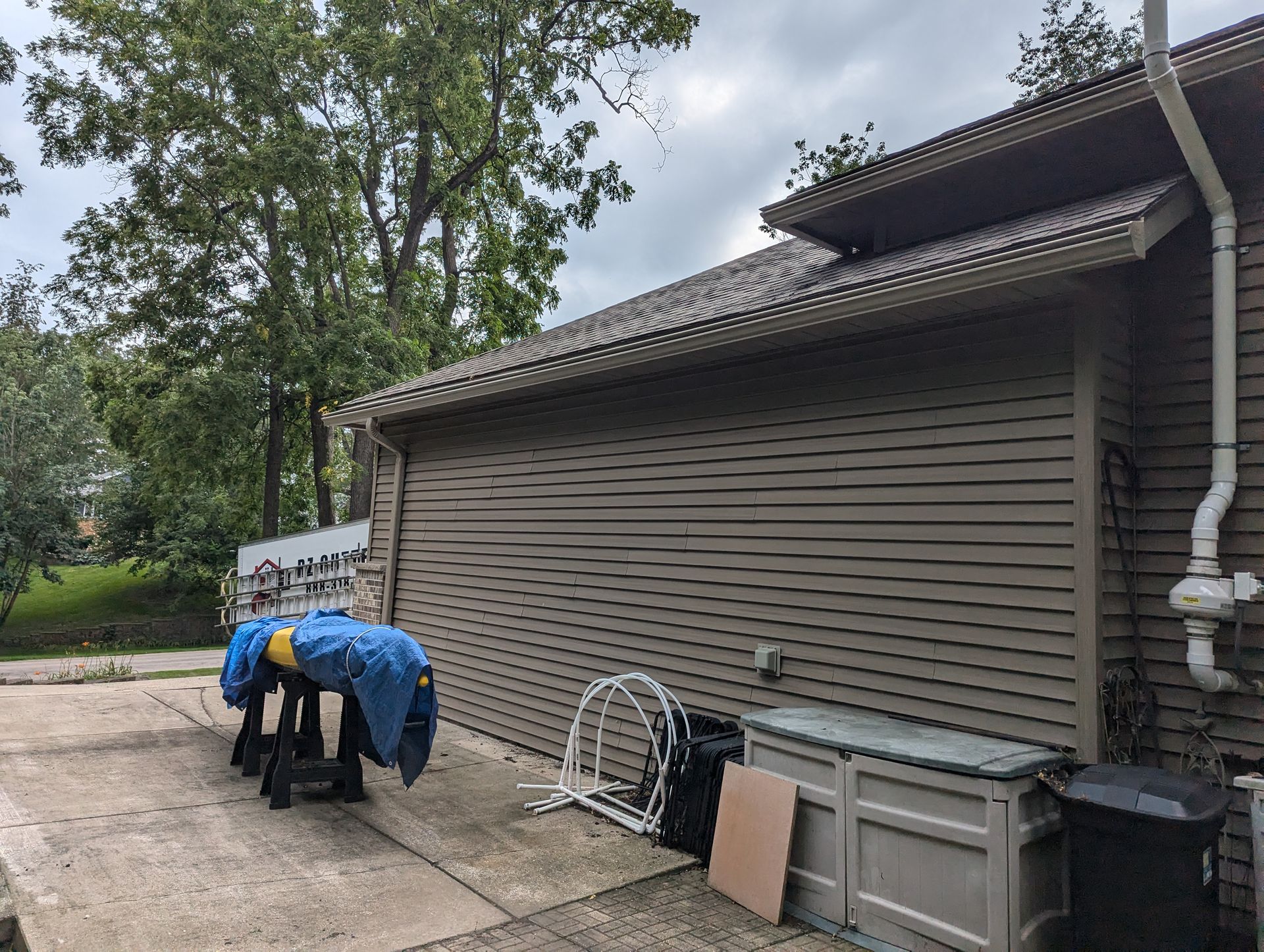 Garage with brown siding, roof, and a covered item on a sawhorse in a driveway.