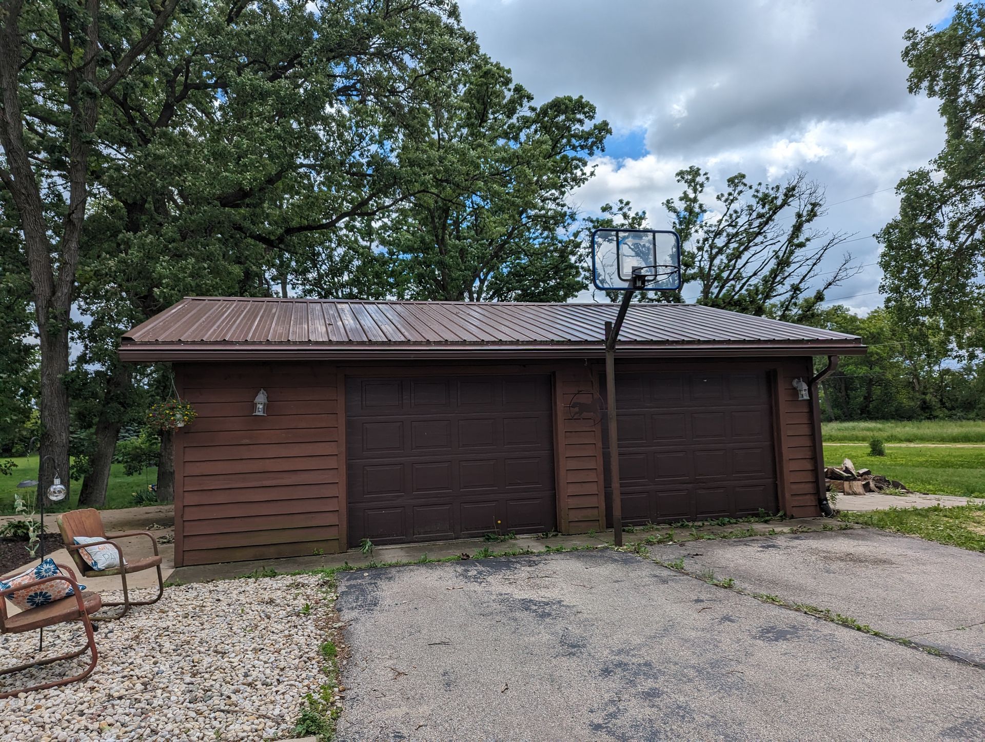 Brown garage with a metal roof, basketball hoop, and driveway. Trees and cloudy sky in background.
