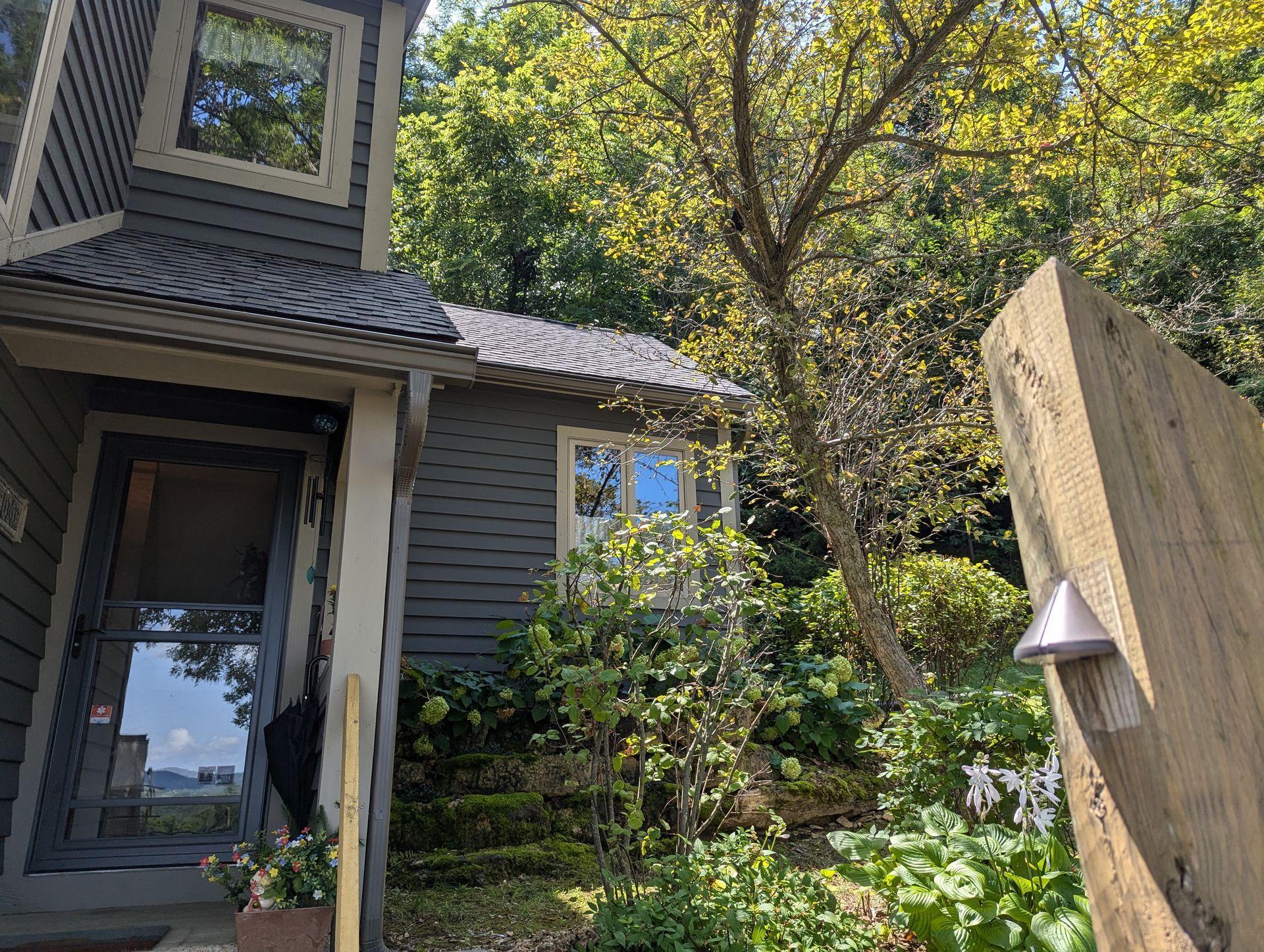 Gray house with a front door, small window, and a wooden post with a light. Surrounded by greenery and trees.