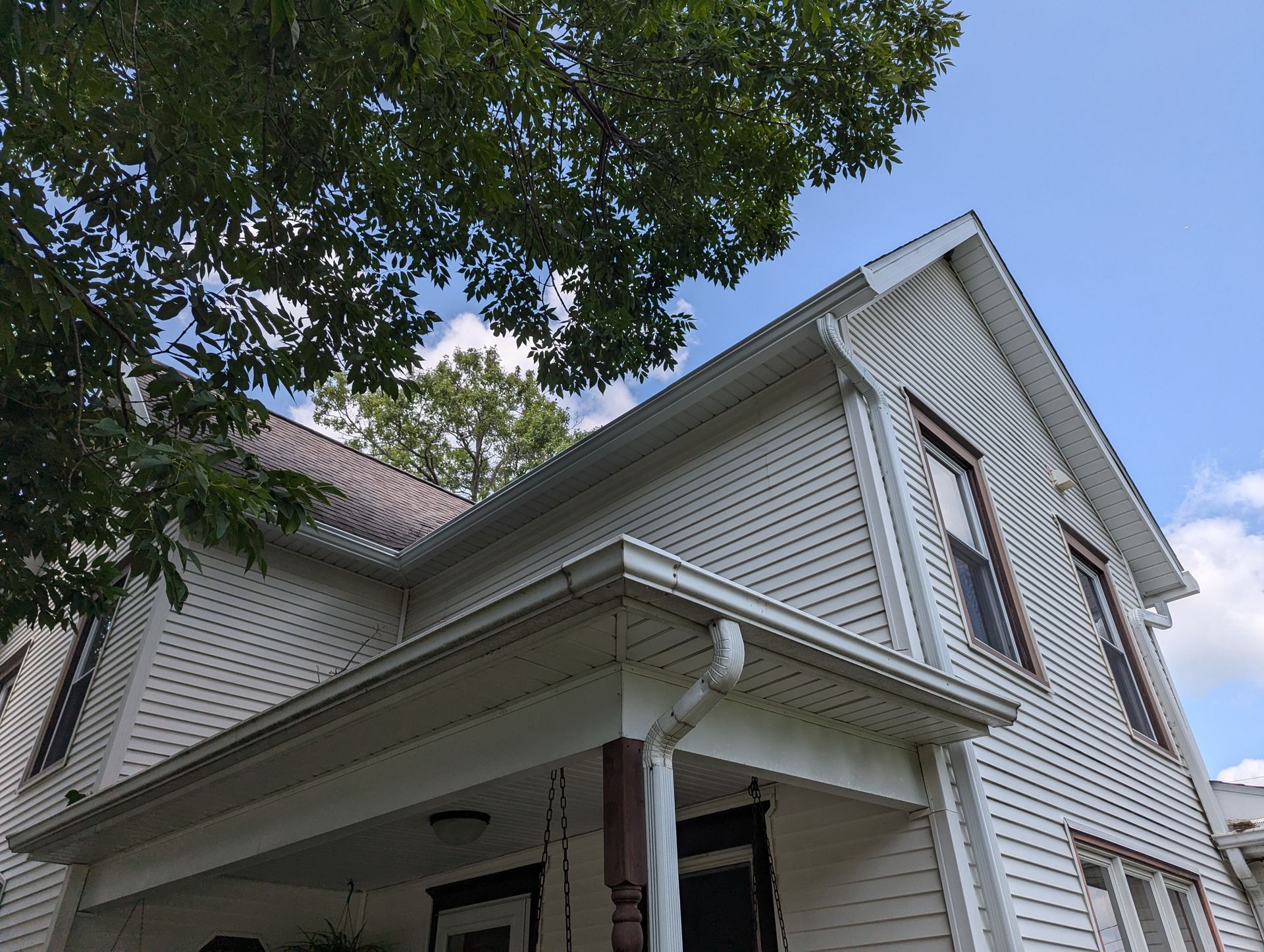 White two-story house with brown trim and gutters under a blue sky, partly obscured by a tree.